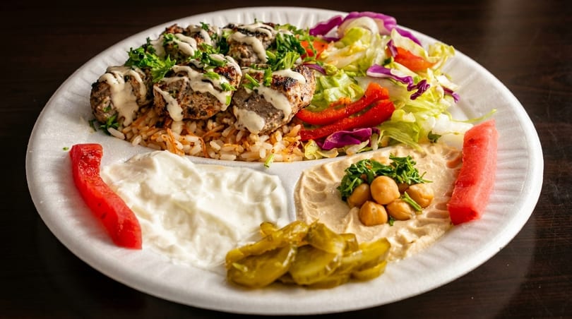 A white plate with kofta, rice, salad, hummus, yogurt, pickles, and watermelon slices.