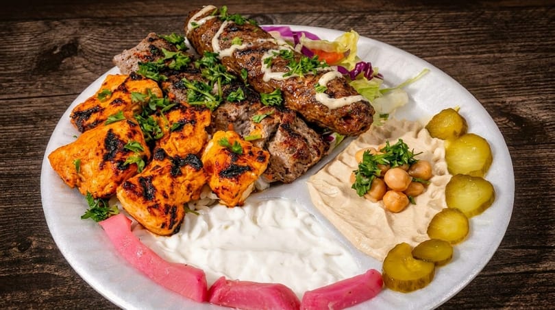 A platter with grilled chicken, beef kebab, hummus, pickles, and a white sauce.