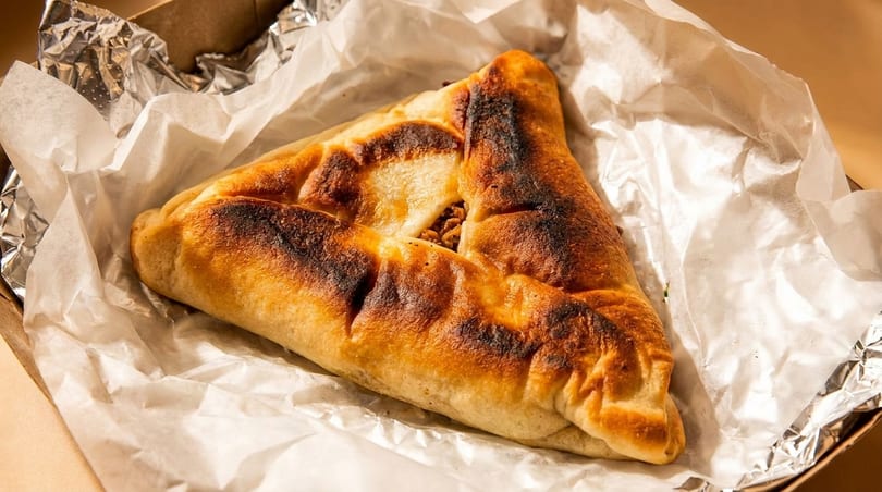 A golden-brown, triangular savory pastry with a meat filling, resting on white parchment paper.