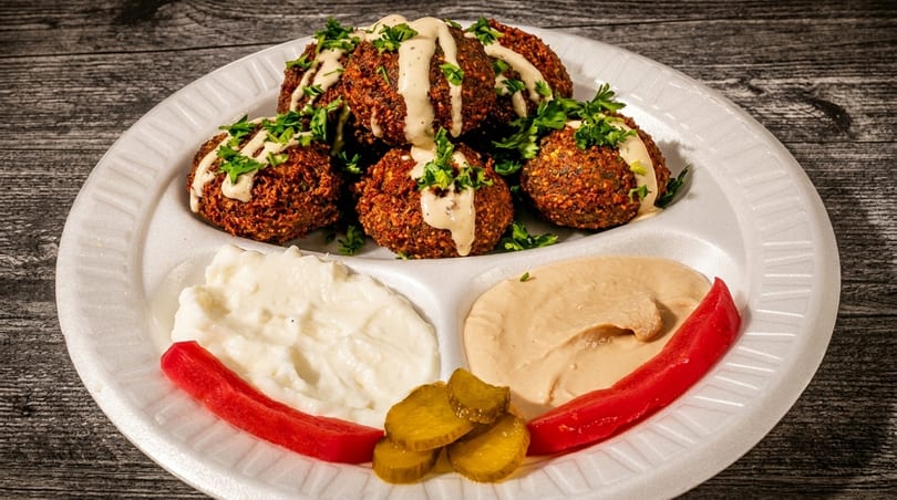 Falafel plate with hummus, white sauce, pickles, and red peppers on a white plate.