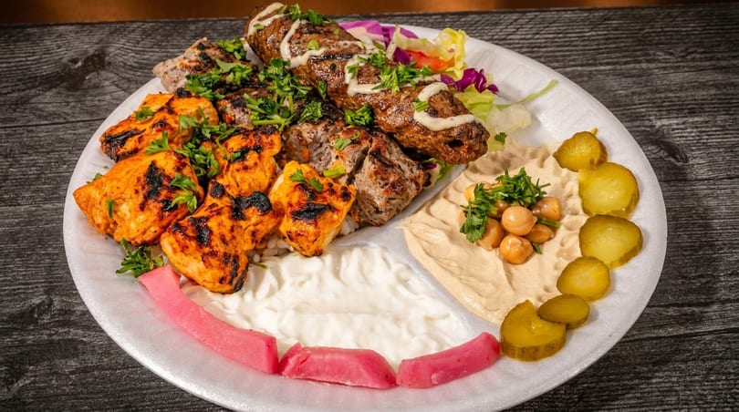 A platter with grilled chicken, beef skewers, hummus, pickles, and salad on a wooden table.