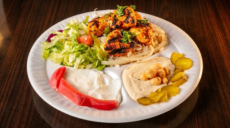 Grilled chicken skewers with rice, salad, hummus, pickles, and white sauce on a white plate.