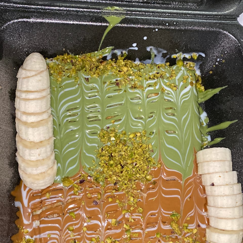 Dessert with green and caramel sauces, white drizzle, chopped pistachios, and sliced bananas in a black container.