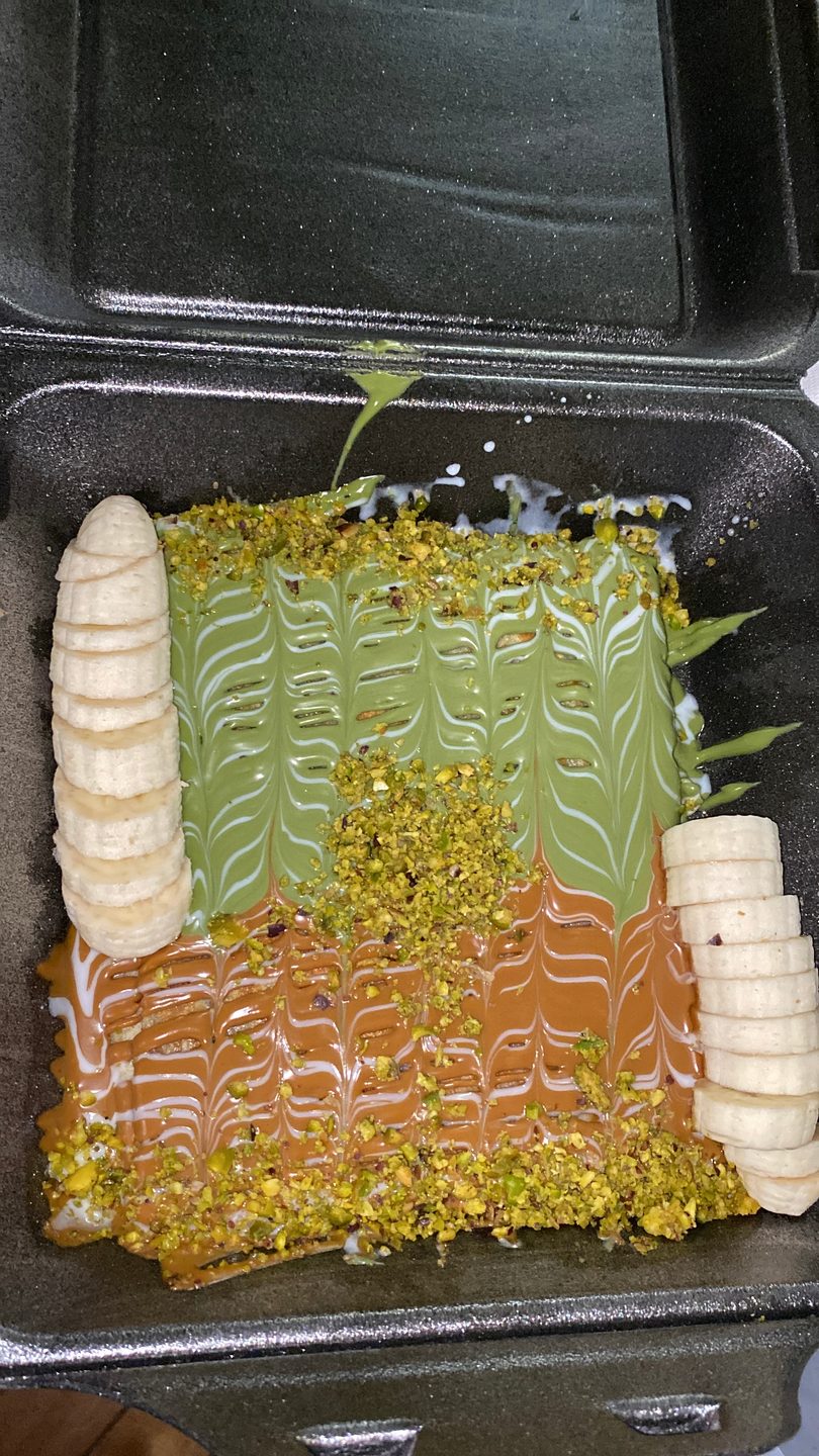 Dessert with green and caramel sauces, white drizzle, chopped pistachios, and sliced bananas in a black container.