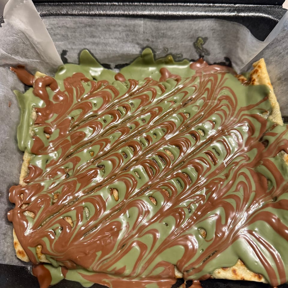 A dessert with green and brown chocolate drizzle in a swirled pattern, served in a black takeout container.