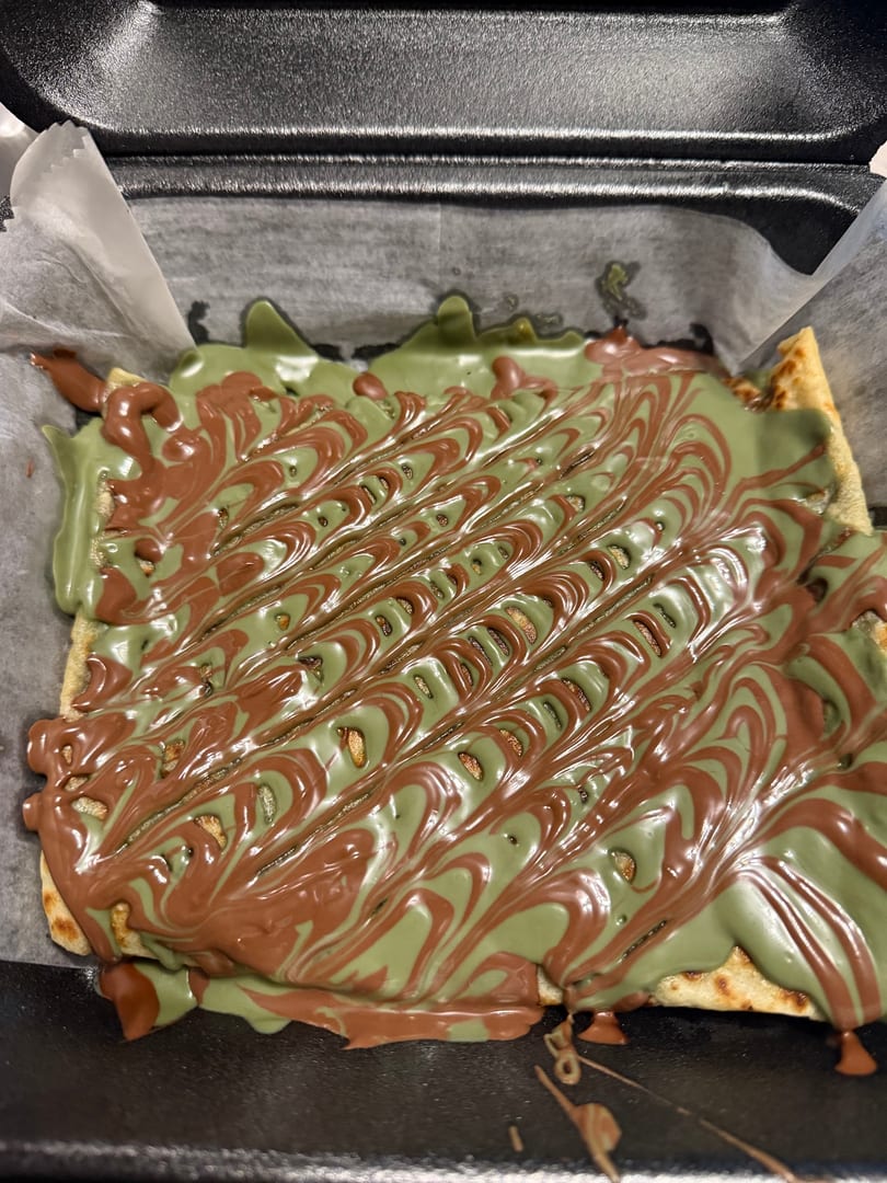 A dessert with green and brown chocolate drizzle in a swirled pattern, served in a black takeout container.